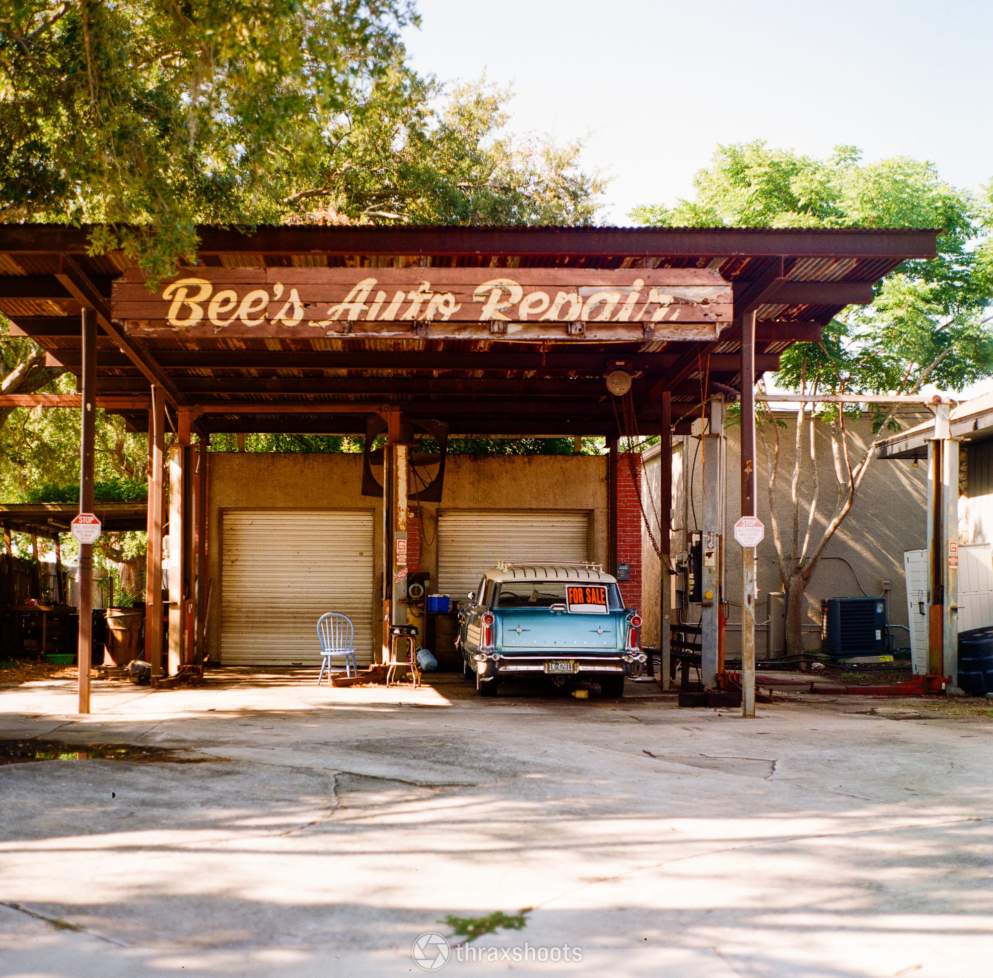 A wide shot of an old, open-air auto shop named "Bee's Auto Repair." The structure has a rustic, corrugated metal roof supported by dark metal pillars, with a faded wooden sign displaying the business name in cursive script. Parked inside the bay is a vintage teal and white station wagon with a red "FOR SALE" sign propped in its rear window. A lone blue plastic chair sits near the closed white garage doors in the background.