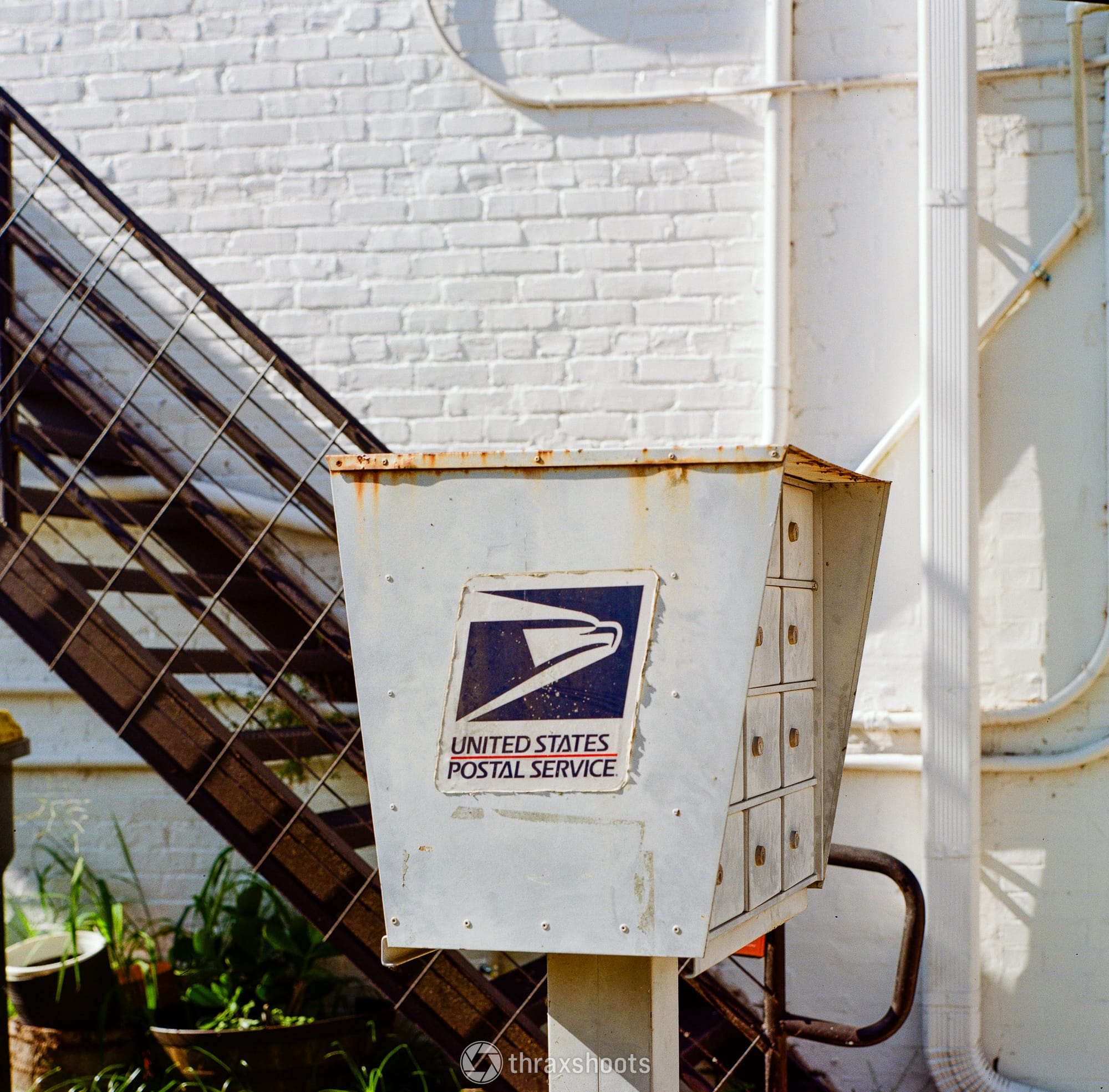 A close-up shot of a weathered, white metal United States Postal Service apartment-style mailbox. The side of the cluster box features a large, slightly faded blue and red USPS eagle logo that reads "UNITED STATES POSTAL SERVICE." The individual keyholes and small metal doors are visible on its side. In the background, an outdoor dark metal staircase runs alongside a white brick wall.