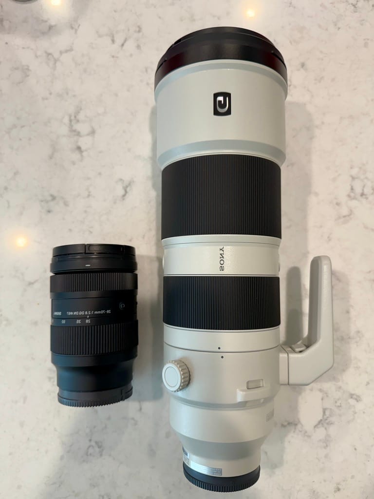 Top-down view of two camera lenses resting side-by-side on a light speckled countertop. On the left is a compact, all-black zoom lens. On the right is a significantly larger, professional white telephoto lens featuring prominent black rubber grip rings and a built-in tripod collar handle.