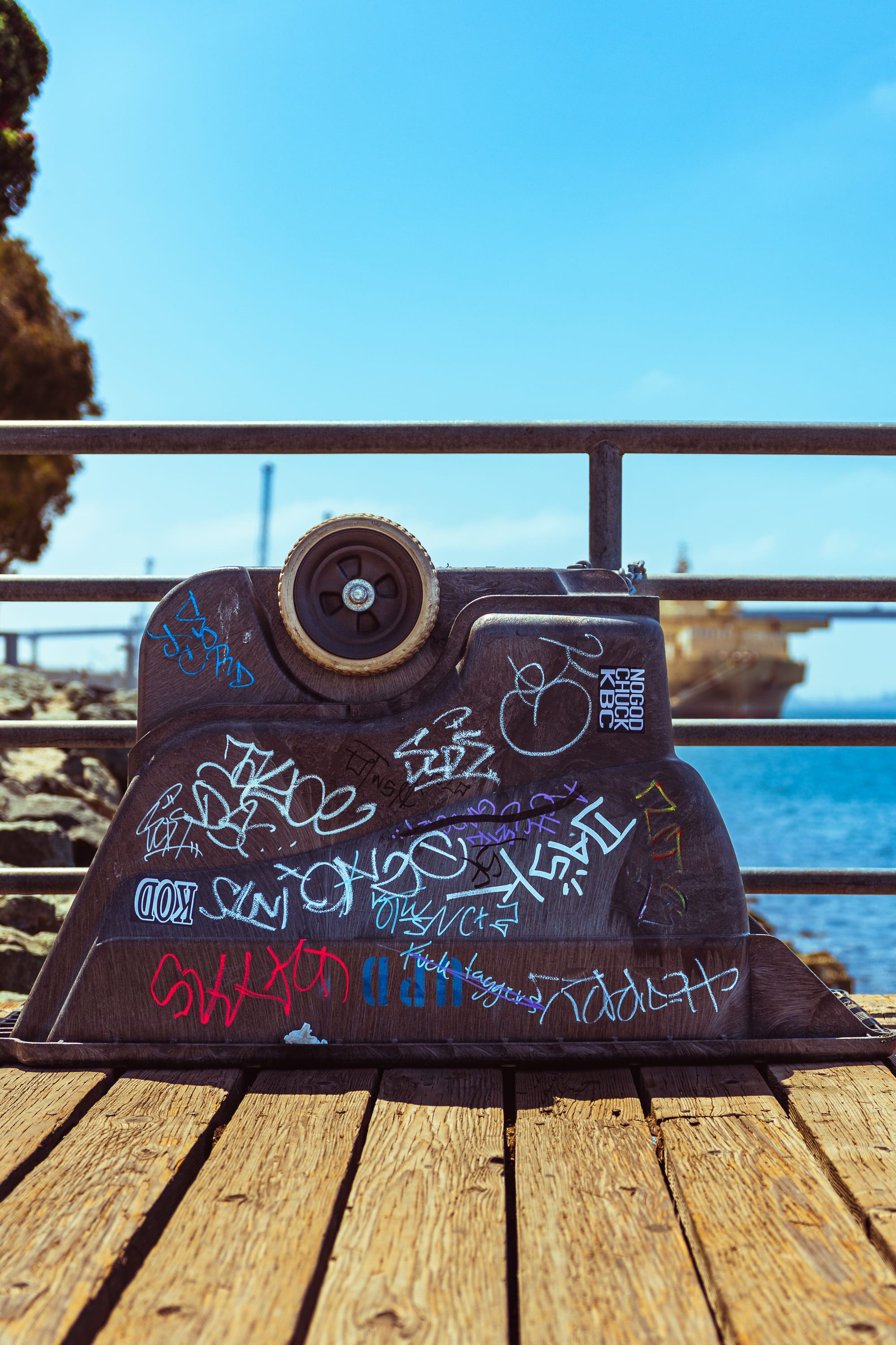 An upside-down black plastic container base covered in white, red, and blue graffiti tags, sitting on a wooden pier boardwalk. In the background, a metal railing overlooks a blue ocean harbor with a blurred large cargo ship under a clear blue sky.