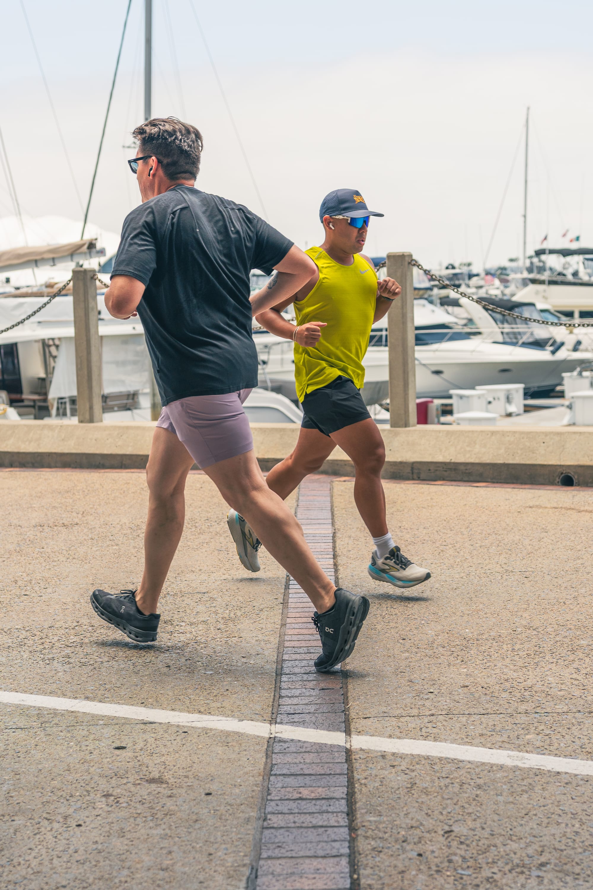 Two men jogging in motion along a paved waterfront path on a bright, slightly hazy day. One man in a black t-shirt and purple shorts leads, followed closely by a man wearing a bright neon yellow tank top, black shorts, sunglasses, and a blue cap. A marina with docked yachts is visible in the background.