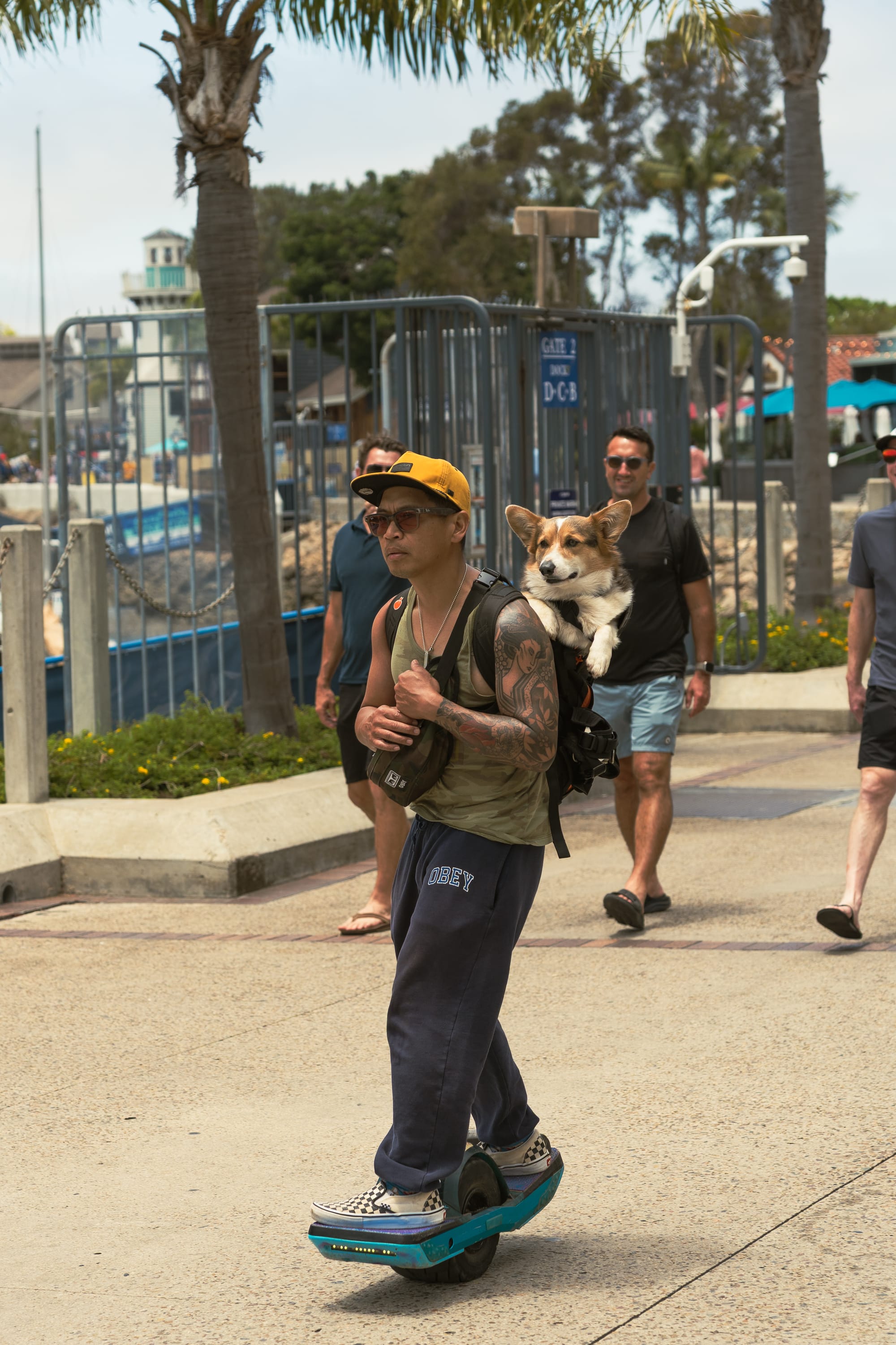 A man with tattoos, wearing a yellow baseball cap, sunglasses, and a green tank top, rides a blue OneWheel skateboard down a paved path. A pembroke welsh corgi dog sits comfortably inside his black backpack, looking out directly at the camera. Other pedestrians walk in the background.