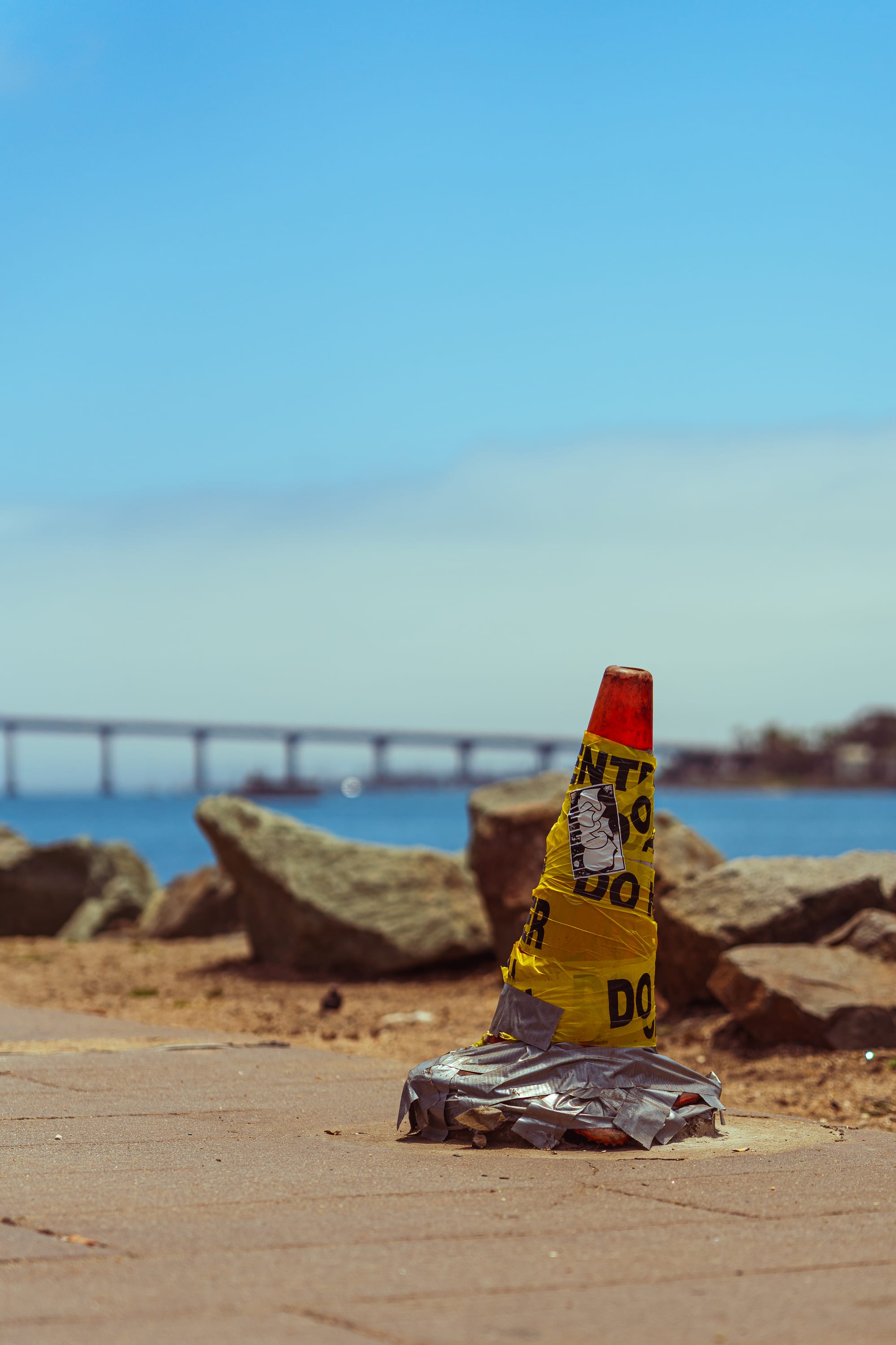 An orange traffic cone heavily wrapped in yellow caution tape and gray duct tape at its base, standing on a paved waterfront path. The background features large gray rocks on a shoreline, a blue body of water, and the distant, sweeping concrete spans of the Coronado Bridge under a light blue sky.