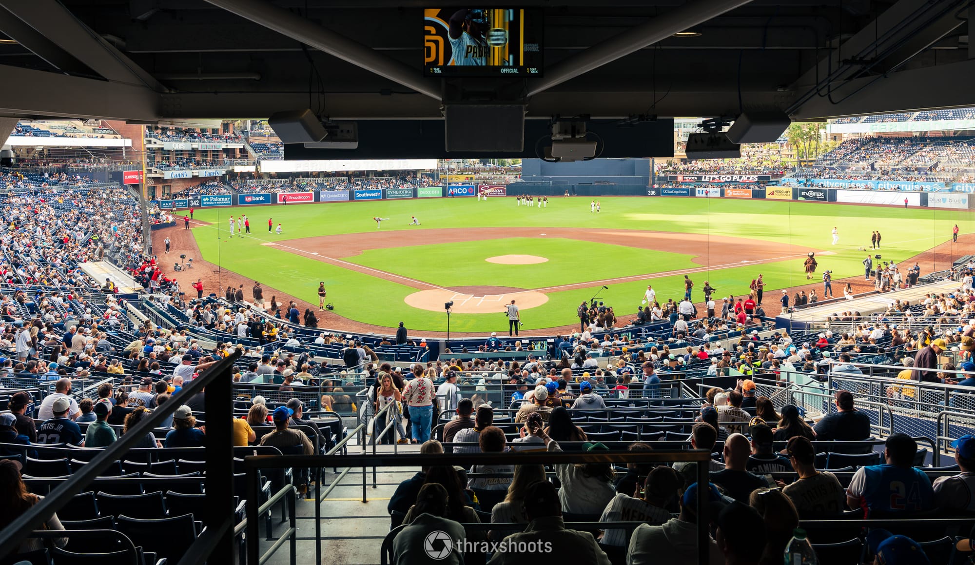 Wide shot from the covered upper stands of a crowded baseball stadium during a daytime game. The baseball diamond, outfield grass, and various advertisements along the wall are visible in the background, while spectators in the foreground sit in tiered seating looking out toward the field.