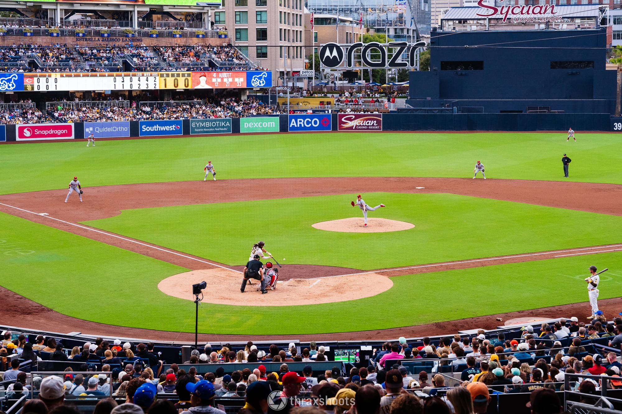 A high-angle, center-field view of a baseball game in progress during the day. A pitcher is mid-throw on the mound, a batter stands ready at home plate with a catcher and umpire behind them, and a full stadium of spectators and advertisements line the background.