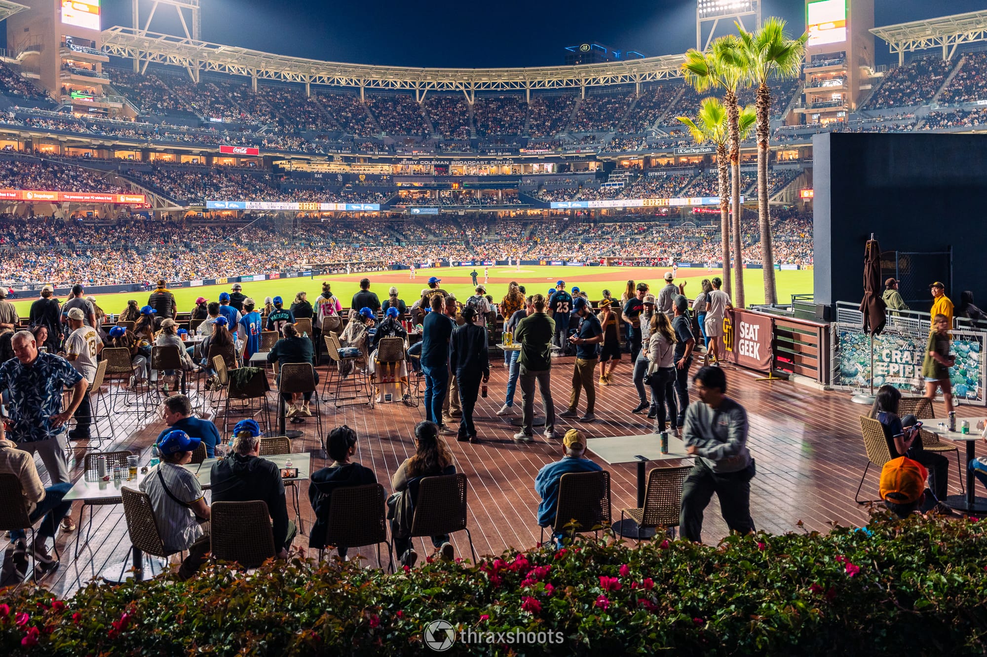 An open-air, multi-tiered outdoor patio or deck area at a baseball stadium at night, featuring fans sitting at tables and standing while socializing. In the background, the brilliantly illuminated stadium stands are filled with people under the bright field lights.