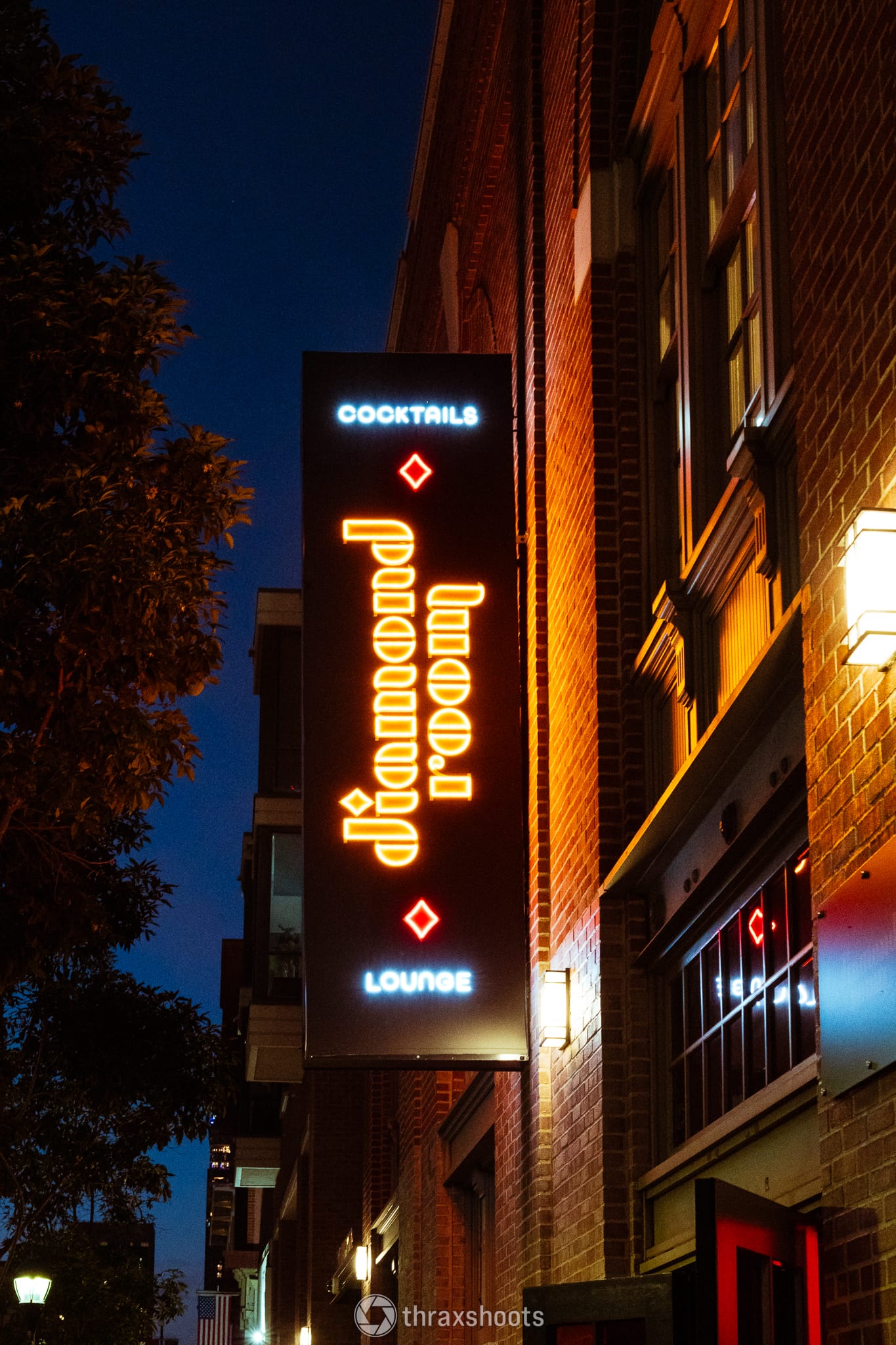 A vertical neon sign attached to a brick building at dusk. The sign features the glowing blue words "COCKTAILS" at the top and "LOUNGE" at the bottom, with a vertically stacked, stylized font in the middle that reads "Diamond Room" lit in warm orange.