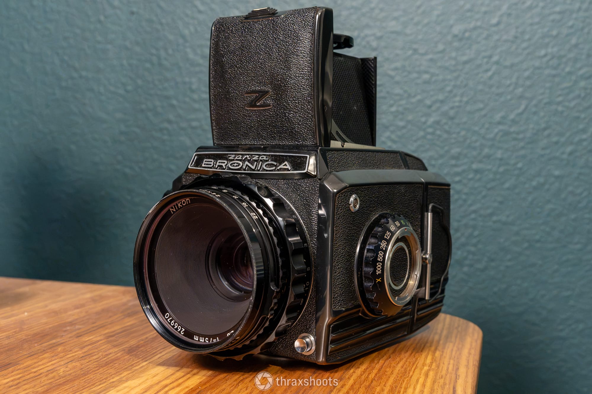 A close-up, front three-quarter view of a vintage Zanza Bronica medium-format film camera resting on a light brown wooden surface. The camera is equipped with a large Nikon 75mm lens facing forward. Its waist-level viewfinder hood is popped open on top, revealing a stylized "Z" logo embossed on the textured front flap. The camera body features a mix of black leatherette panels and metallic dials.