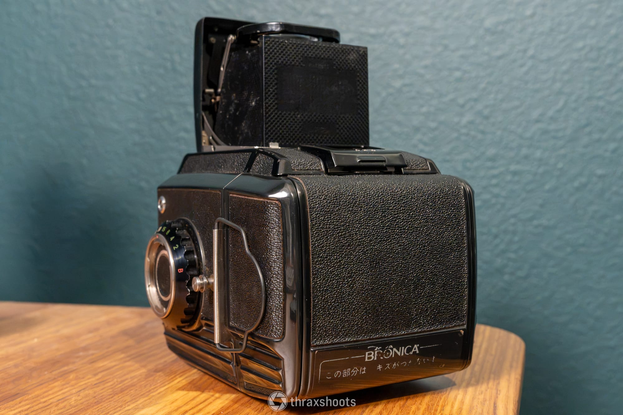 A rear three-quarter view of the black Zanza Bronica medium-format camera on a wooden table. The focus is on the back film magazine, which is covered in a textured black leatherette material. At the bottom of the back panel, the word "BRONICA" is printed next to a line of Japanese text. The open metal mechanism of the waist-level viewfinder is visible from behind on the top of the camera body.